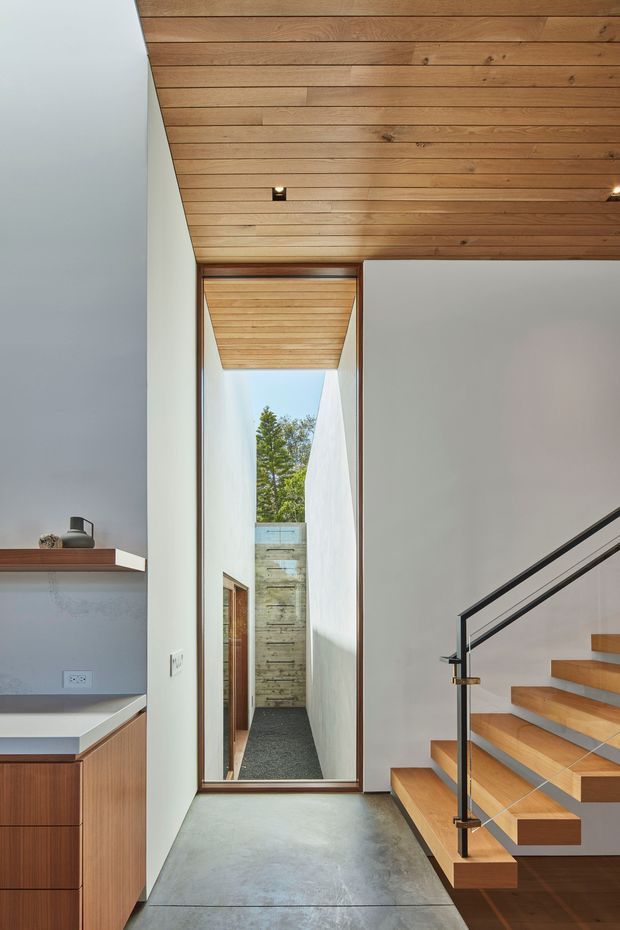 A bright contemporary interior hallway featuring a floating timber staircase with black metal and glass balustrade, a polished concrete floor, white walls, and a warm timber-lined ceiling. A narrow glazed lightwell or courtyard is visible beyond the hallway, while partial built-in cabinetry and a bench appear at left.