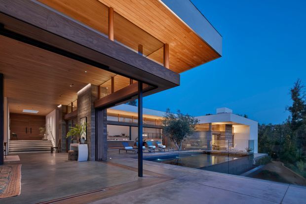 Twilight exterior view of a large contemporary residence organized around a swimming pool and covered outdoor terrace. The home features strong horizontal roof projections, extensive floor-to-ceiling glazing, warm timber soffits and ceilings, dark structural beams and posts, smooth concrete paving, and a minimalist white pavilion-like wing. An open-plan interior and landscaped setting are visible beyond the terrace.