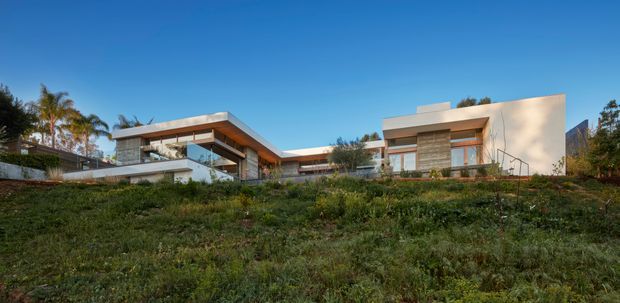 Wide exterior view of a low-slung contemporary residence set on a landscaped, gently sloping site. The home features crisp white rendered volumes, extensive glazing, projecting flat roof planes, timber-toned soffits and screening, and a central connecting section. Palm trees and mature vegetation surround the property under a clear blue sky.