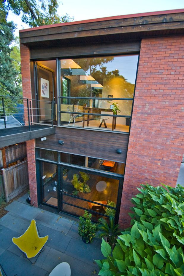 Exterior view of a compact multi-level brick building with dark timber cladding, broad black-framed glazing, and a projecting balcony with slender metal railings. Warmly lit interiors are visible through the windows, while planted greenery and a paved terrace occupy the foreground. The facade combines red brick, timber, and extensive glazing in a contemporary urban residential design.