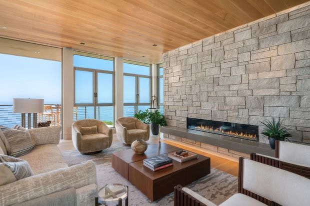 A bright, contemporary living room with expansive floor-to-ceiling glazing overlooking a blue coastal horizon. The room features a full-height grey stone feature wall with a long linear fireplace, warm timber ceiling, pale upholstered seating, sculptural armchairs, a dark timber coffee table, and indoor plants. The space feels refined, warm, and relaxed, with a strong connection to the view.