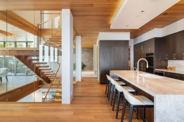 Open-plan contemporary kitchen and circulation space with a large stone island, dark wood cabinetry, timber flooring and extensive timber ceiling lining. A dramatic glazed stairway with timber treads and a glass balustrade overlooks a lower level, while large windows provide abundant natural light.