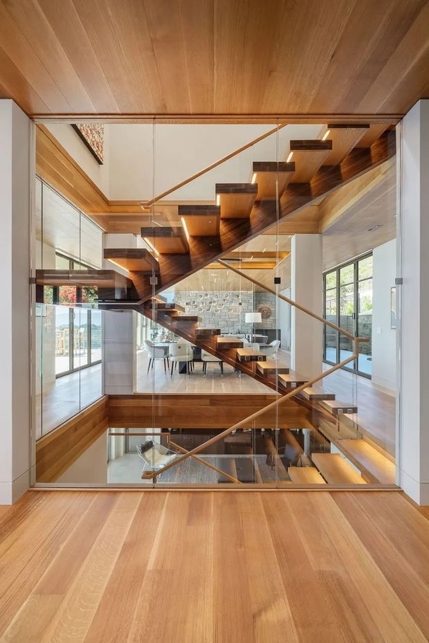 A dramatic interior view centered on a sculptural open-riser timber staircase spanning multiple levels, enclosed by extensive clear glass balustrades and partitions. Warm timber flooring, ceiling, stair treads, and soffits contrast with white walls and bright glazing. A dining area and lower-level living space are visible through the stair void and glass, creating a layered, open-plan composition with strong vertical and diagonal lines.