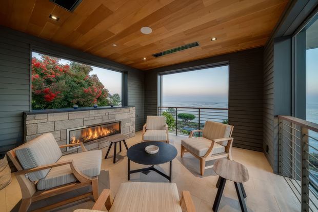 A refined enclosed outdoor lounge or sunroom overlooking the ocean, furnished with light timber-framed lounge chairs and a round coffee table. The space features a built-in stone fireplace with a lit fire, dark horizontal wall cladding, warm timber ceiling boards with recessed lighting, large glazed openings, and a metal cable-and-post balustrade.