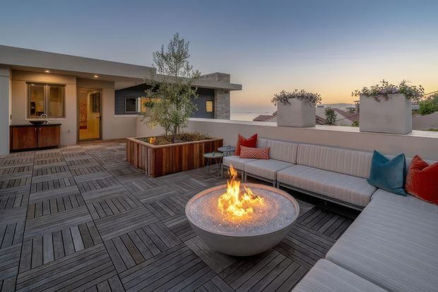 A rooftop terrace at dusk with a broad timber deck, built-in sectional seating, colorful cushions, raised planters, and a central gas fire bowl. A compact modern rooftop structure with an outdoor kitchenette or bar area is visible at the rear, along with distant urban rooftops and the evening sky.