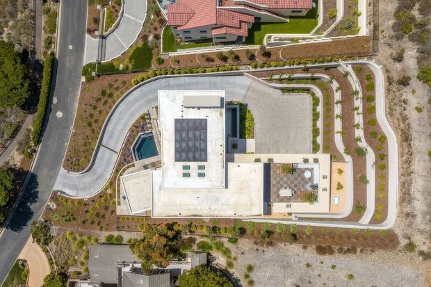 Aerial, top-down view of a contemporary residential property with a large flat-roofed light-colored building, landscaped grounds, curved driveway and paths, a small swimming pool, and a nearby red-tiled-roof house. The image primarily documents the site layout, building footprint, hardscape, and landscape design.