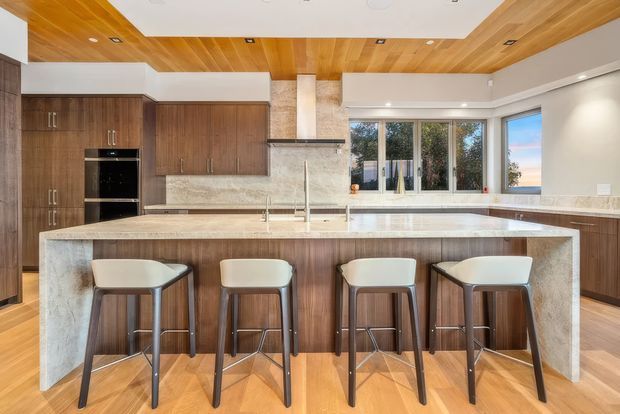 A spacious contemporary kitchen with dark timber cabinetry, a large island with four bar stools, a pale stone benchtop and waterfall ends, and a matching stone splashback. Warm timber lining extends across the ceiling, while large windows provide abundant natural light. Integrated appliances, recessed lighting, and a central rangehood contribute to the refined modern fit-out.