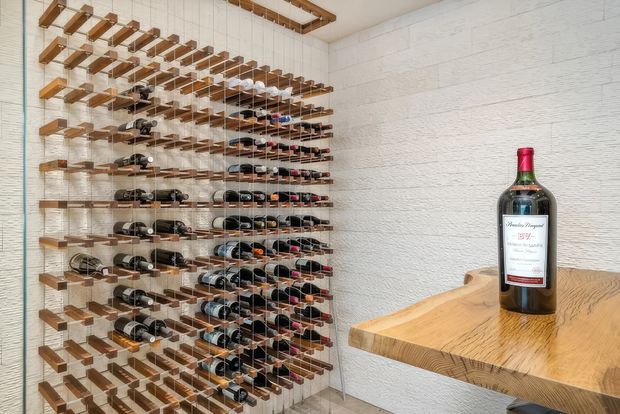 A compact, bright wine storage room featuring a large floor-to-ceiling wine rack made from repeated timber shelves and thin metal supports. A textured white wall and a thick live-edge timber counter or display ledge occupy the right side, with a single wine bottle placed on top. The space has a clean, contemporary cellar or tasting-room appearance.