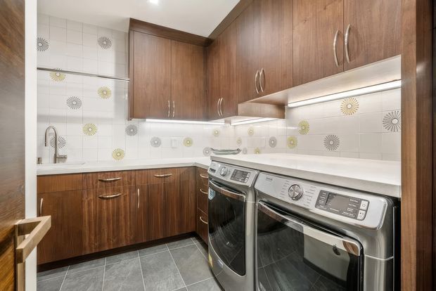 Compact, well-appointed laundry room with extensive warm wood cabinetry, white tiled walls featuring patterned circular accents, white benchtops, under-cabinet lighting, stainless-steel sink and tap, and side-by-side stainless-steel washer and dryer. Grey tiled flooring and integrated storage create a practical contemporary fit-out.