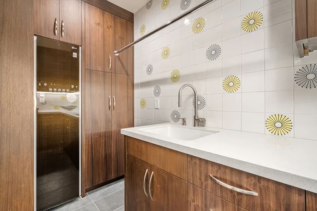 Compact contemporary kitchen with warm wood-grain cabinetry, a white engineered-looking benchtop, integrated appliances, a deep single-bowl sink, and a white tiled splashback decorated with yellow and grey radial floral motifs. Grey floor tiles and stainless-steel hardware complete the practical fit-out.