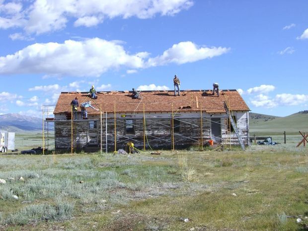 Exterior view of a small rural building under construction or renovation, with workers and extensive scaffolding around the walls and roof. The structure has a simple pitched roof with reddish terracotta-coloured roofing and pale wall cladding or masonry. It sits in an open grassy, mountainous landscape beneath a bright partly cloudy sky.