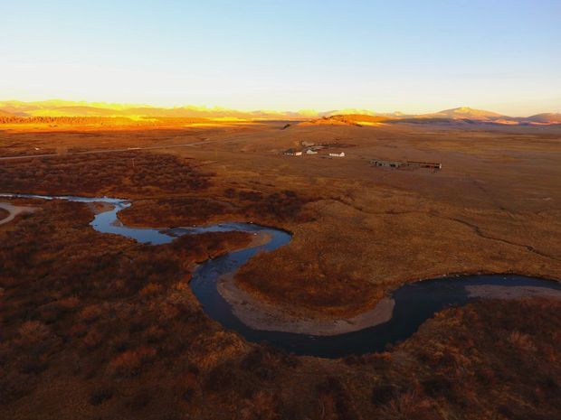 Aerial landscape view of a broad, remote rural plain with a winding dark river or wetland channel in the foreground, low scrub and grasses, scattered small structures, and distant sunlit mountains beneath a clear sky. The scene has warm golden-hour lighting and an expansive, undeveloped character.