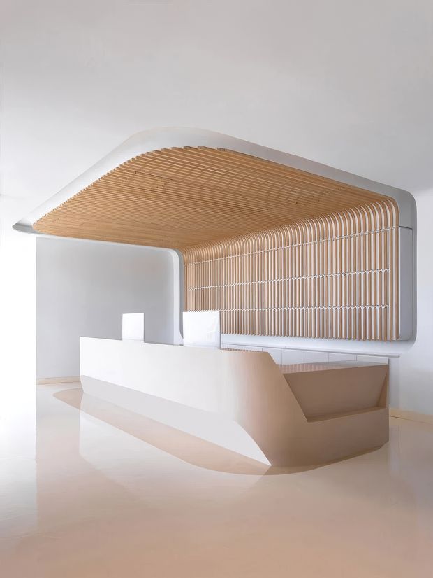 A sculptural, minimalist reception or service counter in a bright contemporary interior. The white curved counter and surrounding wall form a flowing enclosure beneath a rounded canopy lined with closely spaced warm timber slats, creating a soft ribbed feature ceiling and wall. The palette is predominantly white, cream and light timber with smooth seamless surfaces and gentle curves.