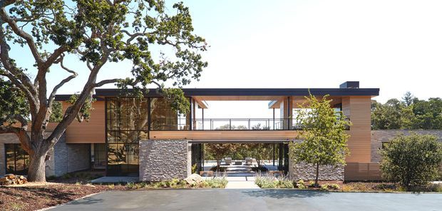 Exterior architectural photograph of a low, modern multi-level residence with a strong horizontal form, broad flat roof, timber-clad upper volumes, exposed structural columns and beams, extensive glazing, and stone-clad lower walls. A central open passage or covered entry creates a framed view through the building, with mature landscaping and a paved approach in front.