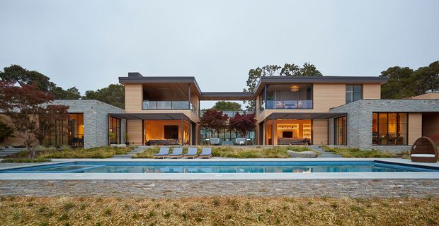 Wide exterior view of a large contemporary residential property arranged as connected pavilion-like wings around a central courtyard and long swimming pool. The low-rise building has flat roof forms, extensive glazing, warm timber-toned wall cladding, dark grey stone masonry, and dark metal window and door frames. Landscaped planting and paved pool surrounds are visible in the foreground.
