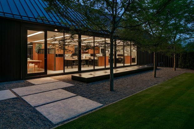 Exterior dusk view of a contemporary single-storey building with dark vertical cladding, a steeply sloped roof, and extensive floor-to-ceiling glazing. The illuminated interior appears to contain a kitchen and dining area. A landscaped courtyard features large concrete stepping slabs, gravel, low rectangular raised planters, and mature trees.