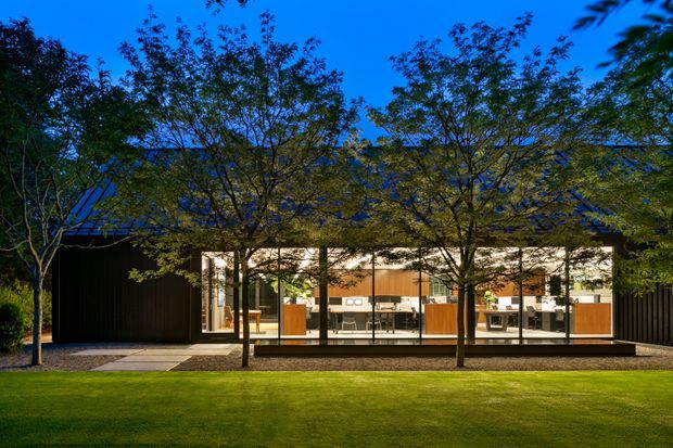 A low, modern pavilion-style building viewed from a landscaped lawn at dusk. The dark ribbed exterior and broad floor-to-ceiling glazed frontage reveal a warmly lit open-plan interior with timber-toned cabinetry and furnishings. Mature slender trees partially screen the facade, and the building has a shallow pitched or gabled roof.