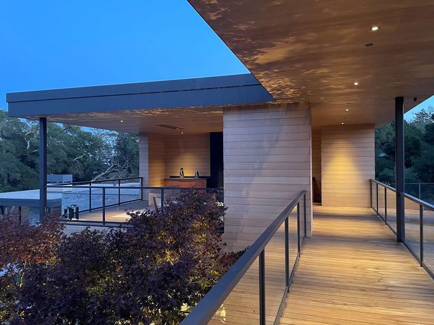 A contemporary elevated outdoor walkway and covered terrace at dusk, with warm timber soffits and decking, slim dark metal posts and railings, and a central light timber-clad wall. The open-sided structure overlooks dense vegetation and incorporates a small sheltered seating or entertaining area. Recessed ceiling lights create a warm, inviting atmosphere.