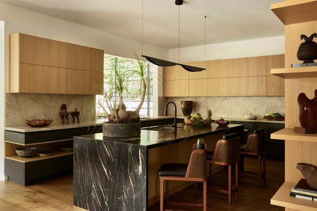 Warm, refined contemporary kitchen with extensive timber cabinetry, a dark veined stone island and benchtops, a matching stone splashback, and an integrated dining or breakfast bar with four upholstered stools. Open shelving displays decorative objects, while a sculptural black pendant light hangs above the island. Large windows bring in natural light and greenery is visible within the space.