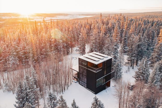 Aerial view of a compact, dark-clad modern cabin set within a dense snow-covered forest. The multi-level structure has a flat roof with raised linear roof elements, large glazed openings, and a projecting deck or balcony. Warm low sunlight filters across the winter landscape, with a broad body of water or open horizon visible beyond the trees.