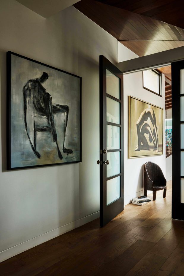 A moody interior view through black-framed glazed doors into an art-filled hallway or entry space. Dark timber flooring continues beneath a dramatic timber-lined angled ceiling, while large monochrome figurative artworks, a dark sculptural chair, and stacked books create a refined gallery-like atmosphere. White plaster walls and daylight from adjacent rooms provide contrast.
