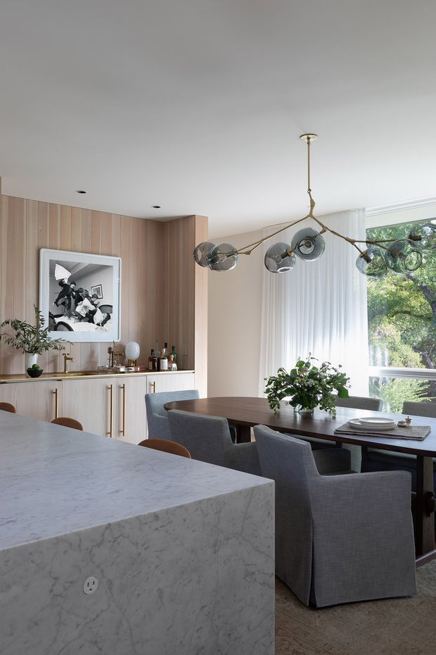A refined open-plan dining and kitchen interior. A long dark wood dining table with upholstered grey armchairs sits beneath a sculptural multi-globe pendant light. The kitchen features pale vertical timber wall panelling, light cabinetry with slim brass pulls, a marble island benchtop, and a large window with vertical sheer blinds overlooking trees. Decorative artwork, bottles, and indoor plants add detail.