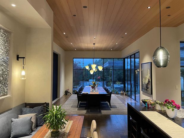 Contemporary open-plan residential interior showing a dining area, living area, and kitchen island beneath a warm timber-lined ceiling. Full-height black-framed glazing opens views to an outdoor area, while pendant lights, dark cabinetry, timber flooring, and neutral plaster walls create a refined, warm atmosphere.