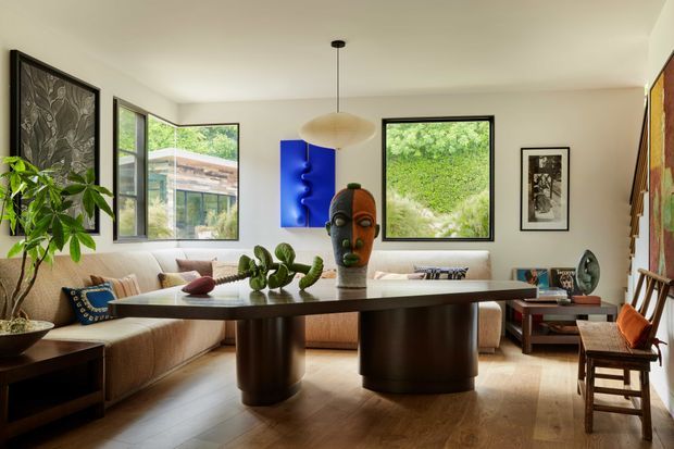 A warm, art-filled contemporary living room with a large sectional sofa, sculptural dark wood or stained timber coffee table, and a central sculptural artwork. The room has pale walls, timber flooring, expansive black-framed windows overlooking greenery, a linear pendant light, framed artwork, indoor plants, and a relaxed eclectic arrangement of furniture and objects.