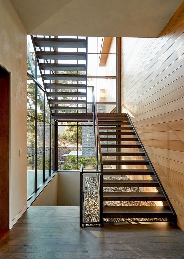 A bright, modern interior stairway with two open-riser flights forming a partial stairwell bridge. Dark metal stringers and railings contrast with timber treads, a warm horizontal timber-clad wall, and large floor-to-ceiling glazing. A landscaped pebble-filled courtyard is visible beyond the glass, while dark timber flooring continues through the landing and hall.
