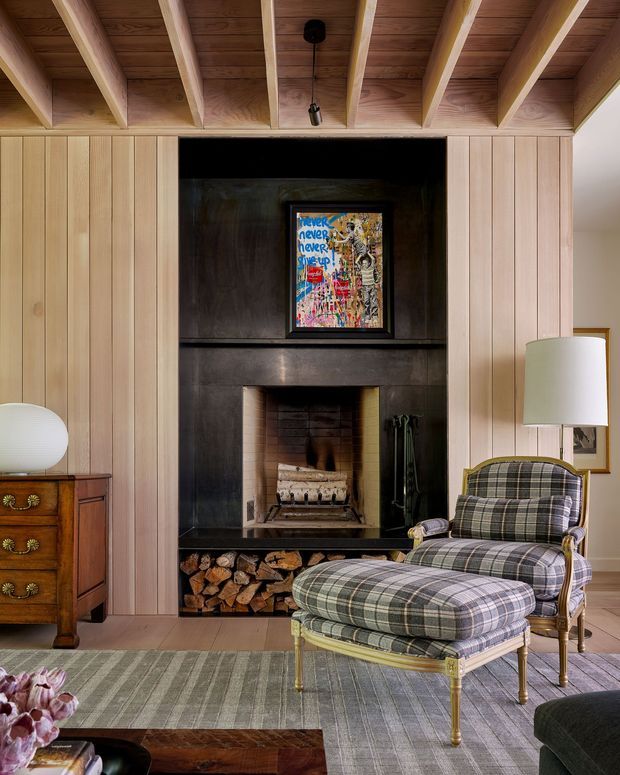 A warm, refined living room centred on a dark, built-in fireplace with a timber-lined surround and stacked firewood storage below. Vertical light timber wall panelling and exposed timber ceiling rafters frame the fireplace, while a plaid upholstered armchair and footstool sit on a large muted area rug. Traditional timber furniture, a floor lamp, framed artwork and soft neutral finishes complete the inviting interior.