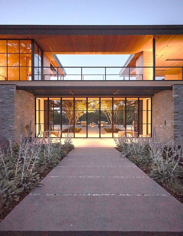 A dusk exterior view of a contemporary two-storey residence, approached along a paved garden path. The facade combines extensive black-framed floor-to-ceiling glazing, warm timber soffits and ceilings, and horizontally coursed grey stone walls. A glazed upper-level terrace or bridge creates a strong open void between two wings, while landscaped planting frames the entry.
