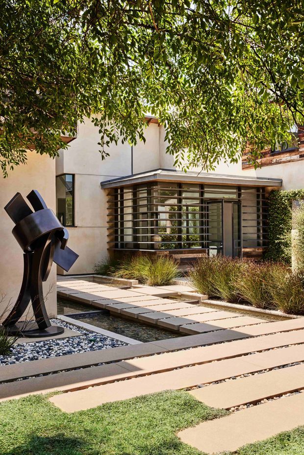 Exterior view of a contemporary low-rise residence framed by mature leafy trees. The entry courtyard features broad pale concrete stepping slabs separated by dark gravel channels, a shallow linear water feature, clipped shrubs and ornamental grasses. A dark abstract metal sculpture stands in a pebble bed beside the path. The building has pale rendered walls, extensive horizontal glazing with dark metal framing and horizontal sun-shading bars, and a low projecting roof.
