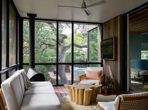 A warm, nature-connected living room with extensive black-framed glazing overlooking mature trees and a swimming pool. The room features a white sofa, sculptural white freestanding fireplace, timber-panelled feature wall with a mounted television, rattan armchairs, a sculptural natural timber coffee table, and a ceiling fan. A bedroom or adjacent room is visible through the open doorway on the right.