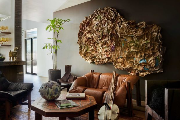 A sophisticated living room with a large abstract sculptural wall artwork as the focal point, a cognac leather sofa and black leather seating arranged around a low octagonal timber coffee table. The room includes a dark fireplace or built-in feature at right, a tall potted indoor tree, sculptural objects, and bright floor-to-ceiling glazing at the rear. The palette is warm and earthy with strong textural contrast.