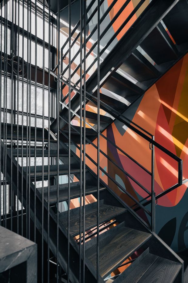 A dramatic interior view of a multi-level industrial-style stairway. Open-riser stairs with dark timber treads and black metal stringers, railings, and dense vertical balustrade rods rise through a tall glazed void. A bold geometric mural in orange, red, navy, and yellow covers the adjacent wall, with strong sunlight creating angular shadows.