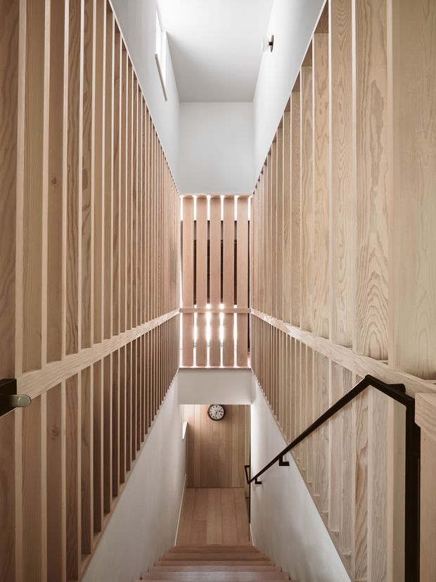 A narrow, double-height stairway with a straight flight of timber stairs descending to a lower landing. Pale timber slat screens and vertical timber wall panelling line both sides, creating a rhythmic architectural enclosure, while white plaster walls and ceiling keep the space bright. A dark metal handrail contrasts with the light wood, and a small round wall clock is visible at the lower level.