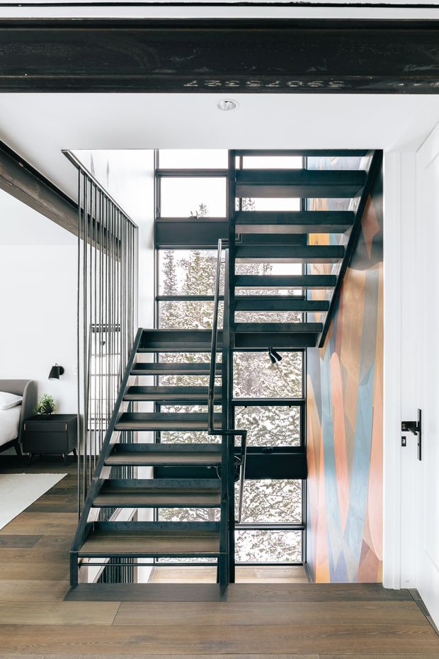 Interior architectural photograph focused on a contemporary open-riser staircase with dark metal stringers, treads, and slender vertical balustrade rods. The stair rises beside expansive floor-to-ceiling glazing, with a colourful geometric feature wall alongside it. Warm timber flooring, white walls and ceiling, and a small portion of an adjacent living area are visible. The space is bright, refined, and vertically open.
