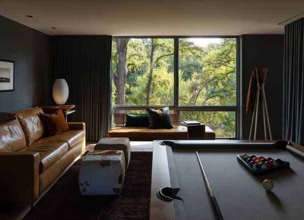 A moody, refined living room with a large billiards table in the foreground, a cognac leather sofa, dark textured wall and curtain treatments, and a built-in window seat overlooking dense greenery. The room combines warm timber and leather tones with charcoal architectural finishes and abundant natural light through expansive glazing.