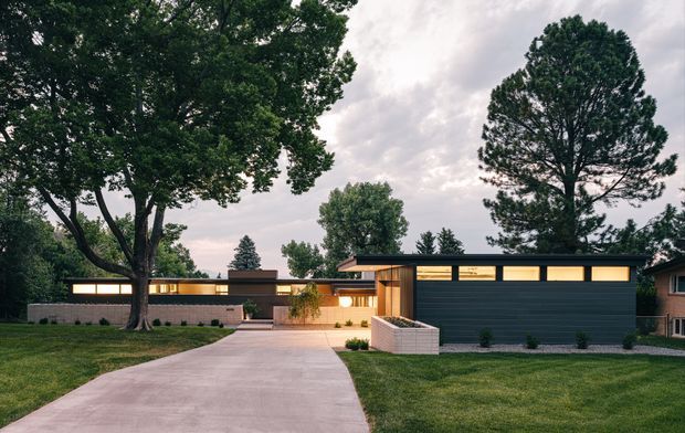 A low, single-storey modern residence viewed from the street at dusk, with a broad concrete driveway leading to a dark horizontal-panel garage or frontage. Warmly illuminated clerestory windows and timber-toned entry elements contrast with the dark facade, while mature trees and lawns frame the property.