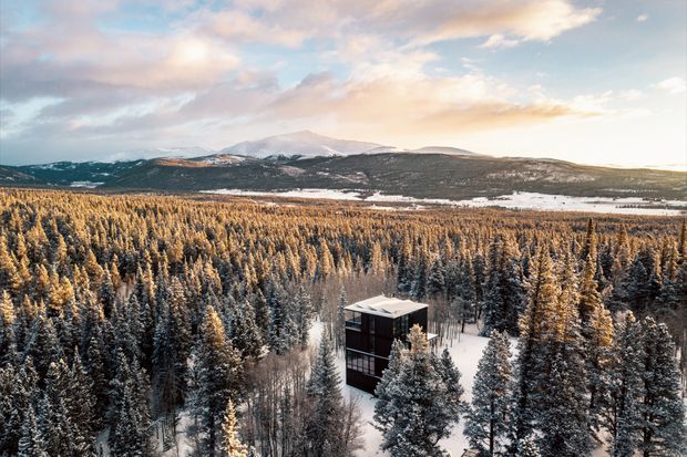 Aerial view of a compact multi-storey dark-clad building nestled among snow-covered evergreen forest, with expansive mountainous terrain and a partly snowy landscape beyond. The scene has a secluded alpine setting and warm low-angle light beneath a partly cloudy sky.