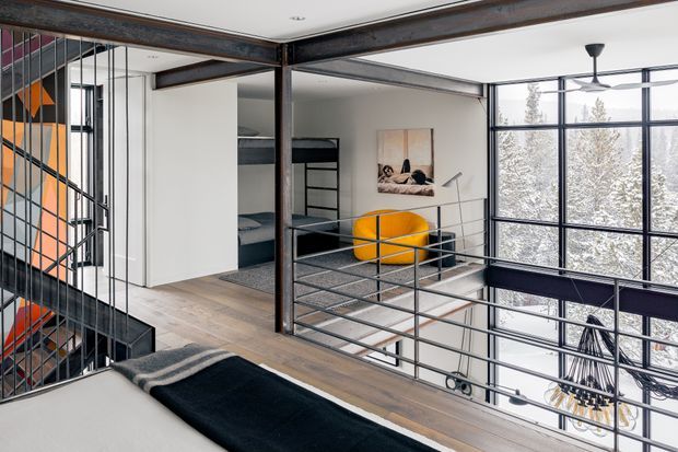 A contemporary industrial-style multi-level interior with an open mezzanine overlooking a lower living area. Black steel beams, posts, railings and stair structure contrast with white walls and pale timber flooring. The space includes an upper sleeping area in the foreground, bunk-style built-in beds behind a partial partition, a bright yellow lounge chair, large black-framed windows, and a ceiling fan. A sculptural orange-and-black wall feature is visible beside the stairs, with trees seen outside through the glazing.