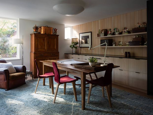 A warm mid-century styled home office and dining workspace with a substantial timber table, upholstered dining chairs, a leather lounge chair, and extensive built-in timber wall cabinetry and shelving. The room is naturally lit through large windows, with a textured blue-grey carpet, wood-panelled joinery, desk lamps, books, plants, and collected decorative objects. A sculptural flush-mount ceiling light and vintage furniture create a refined, cozy atmosphere.