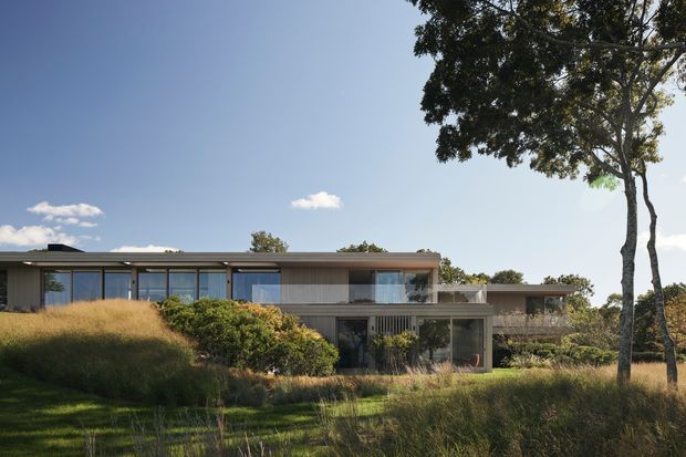 Exterior view of a low-profile contemporary house set within a landscaped, grass-covered site. The home has elongated horizontal proportions, extensive floor-to-ceiling glazing, flat roof planes, dark slender window frames, and a mix of light vertical cladding and darker solid wall sections. Mature trees and dense planting surround the building.