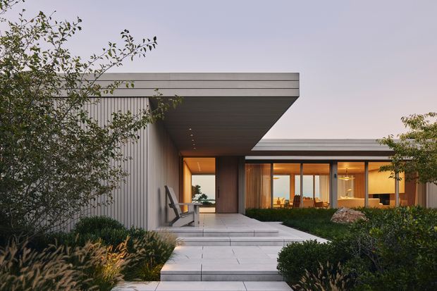 A low, contemporary single-storey residence viewed from a landscaped front approach. The home features strong horizontal roof planes, a deep cantilevered entry canopy, vertical ribbed exterior cladding, expansive warm-lit glazing, and a broad paved path framed by dense planting.
