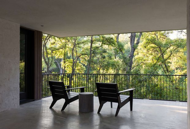 A covered outdoor terrace or balcony overlooking dense woodland, with two dark timber lounge chairs and a small cylindrical side table on a smooth concrete floor. The space has a low flat ceiling, black metal balustrade, deep overhang, and a large dark-framed opening at the left. The setting feels restrained, modern, and connected to nature.