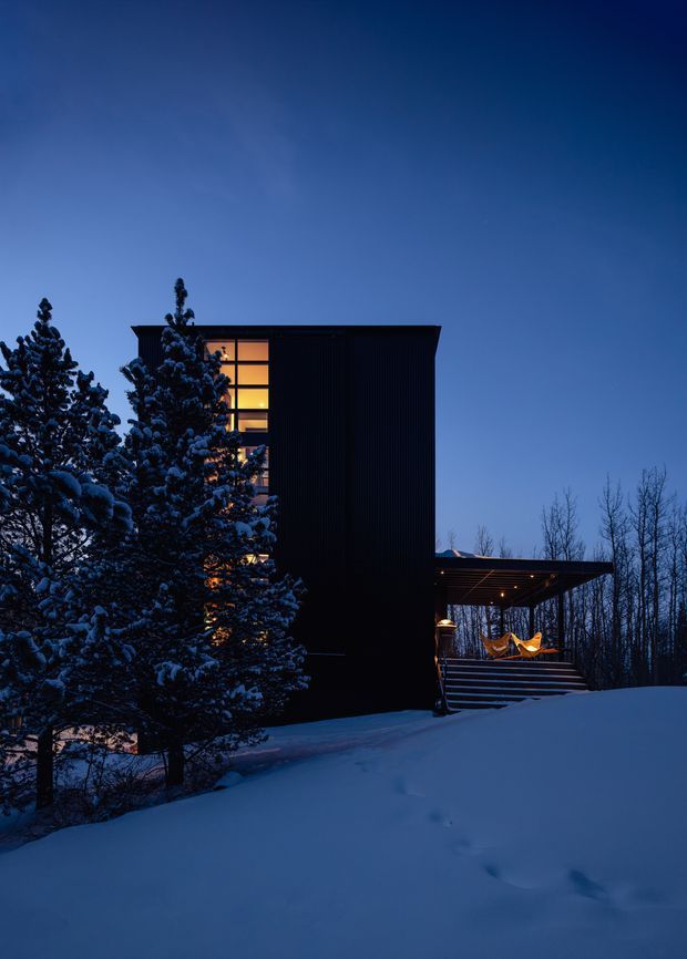 Twilight exterior view of a dark, rectilinear multi-storey house set in a snowy, wooded landscape. Warm light glows through tall vertical windows, while a covered elevated entry or deck with stairs and illuminated seating projects to the right. Snow-covered evergreen trees partially screen the facade, creating a dramatic contrast between the cool blue surroundings and warm interior lighting.