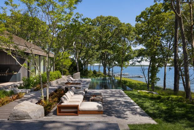 A landscaped waterfront outdoor living area with a long rectangular swimming pool, timber-framed lounge seating, stone-paved terraces, and a low modern pavilion partially visible to the left. Mature trees frame views across the water, while layered stone retaining elements and planting create a natural, refined setting.
