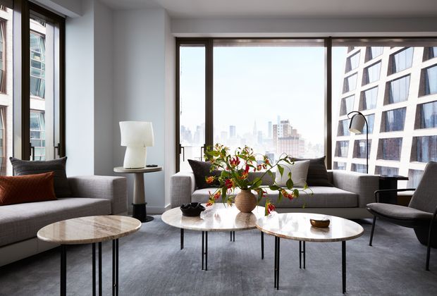 Bright contemporary living room in a high-rise urban apartment, featuring floor-to-ceiling corner glazing, expansive city views, grey upholstered sofas, nested round marble-look coffee tables, a black arc floor lamp, and indoor plants. The room has a refined neutral palette with strong natural light and modern furniture.
