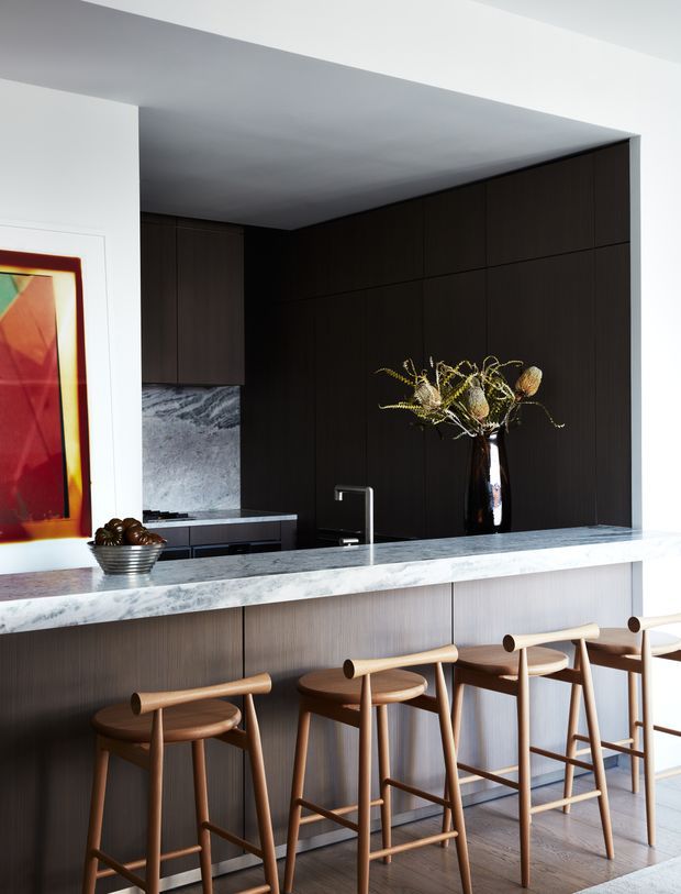 A refined contemporary kitchen with a long island and bar seating. Dark timber cabinetry forms a dramatic full-height feature wall, contrasted with a pale marble-look stone benchtop and lighter wood-grain island cabinetry. A slim black-and-metal tap, integrated sink, abstract artwork, and sculptural dried floral arrangement add visual accents. Several timber bar stools line the island, and timber flooring continues through the space.