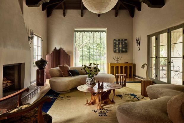 A richly styled living room with a high exposed timber-beam ceiling, plastered walls, large leaded-glass windows and French doors, and a substantial masonry fireplace. Curved upholstered sofas surround a sculptural wood coffee table on a large patterned rug, with a warm timber console and decorative lighting. The room has an eclectic, luxurious and slightly rustic character.